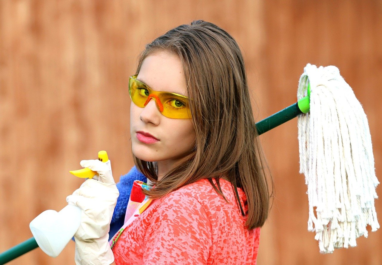 An image of a young woman ready for deep cleaning, wearing yellow protective goggles and white gloves while holding a spray bottle and carrying a mop, perfectly illustrating essential spring cleaning safety tips.