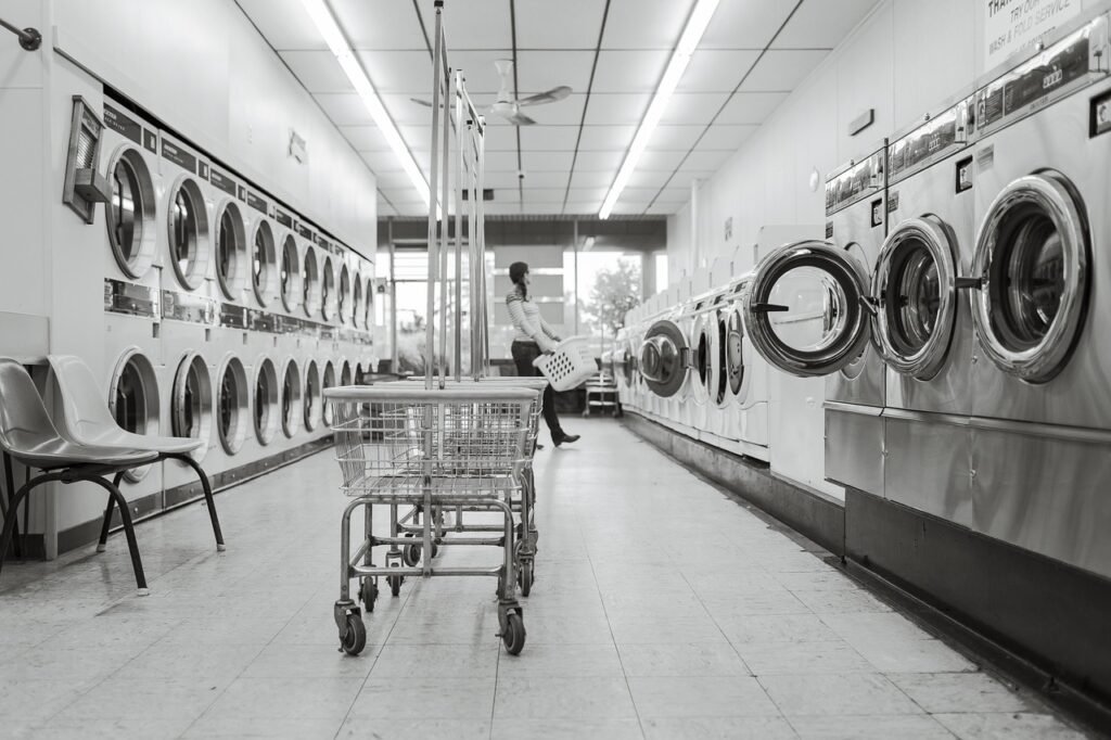 A black and white wide-angle shot of a clean, organized laundromat with rows of industrial washing machines and laundry carts, illustrating the organized outcome of spring cleaning safety tips in a laundry routine.